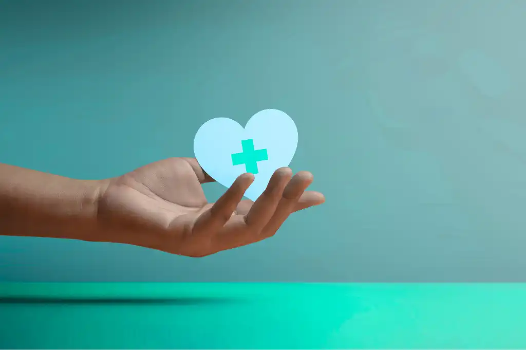 Hand holding a blue heart icon with a medical cross symbol on a teal background.